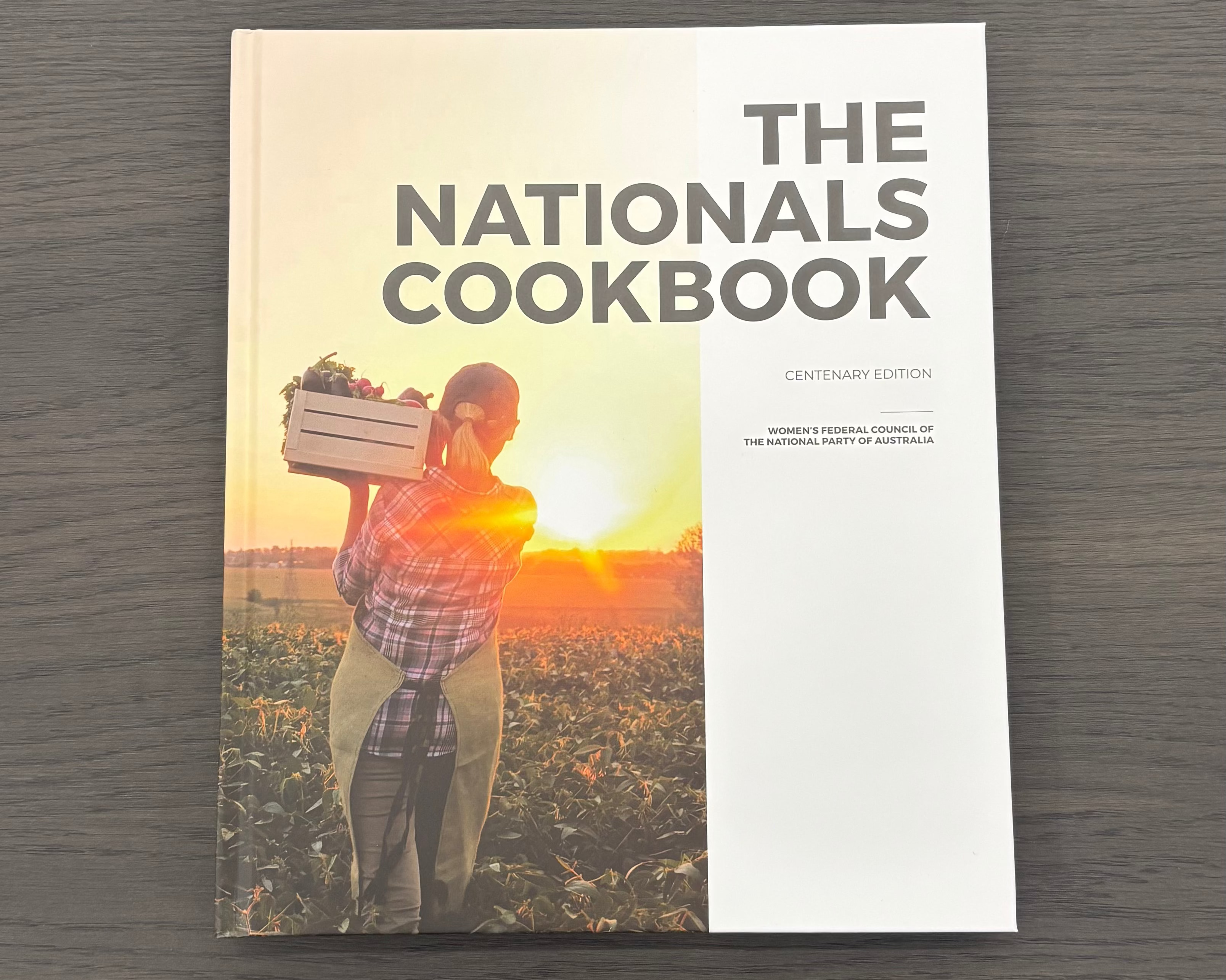 The Nationals Cookbook Centenary Edition book lying flat on a dark gray wood-grain surface. The cover features a split design with a photographic scene of a person harvesting in a field at sunset on the left, and a solid white panel on the right with the title in bold gray text.